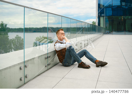 Thoughtful middle-aged man sitting on floor near glass railing beside river on cloudy day Thoughtful middle-aged man sitting on floor near glass railing beside river on cloudy day 140477786