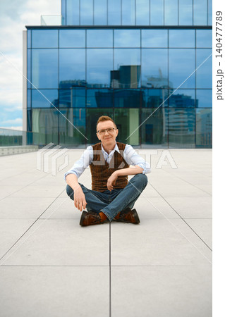 Cheerful middle-aged man sits cross-legged on floor of outdoor terrace at daytime Cheerful middle-aged man sits cross-legged on floor of outdoor terrace at daytime 140477789