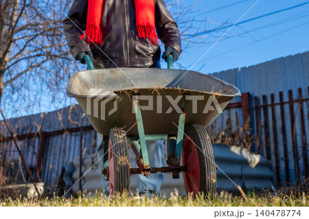 Man gardener pushing wheelbarrow in yard during winter. Male worker using equipment for yard cleaning. Concept of home maintenance, garden preparation, hard labor and seasonal outdoor work. 140478774