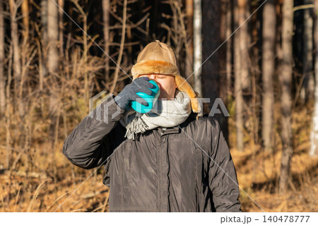 Man wearing winter hat and scarf drinking hot beverage from travel mug in autumn forest. Concept of cozy hiking and outdoor leisure activity during cold weather in nature landscape. Man wearing winter hat and scarf drinking hot beverage from travel mug in autumn forest. Concept of cozy hiking and outdoor leisure activity during cold weather in nature landscape. 140478777