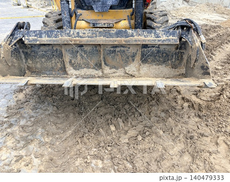 A sturdy metal construction bucket rests on a mound of loose, lumpy soil and sand. Construction area A sturdy metal construction bucket rests on a mound of loose, lumpy soil and sand. Construction area 140479333