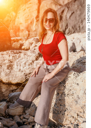 Woman rocks seaside Smiling middle aged woman in red top sits on coastal rocks during a golden sunset at the beach 140479669