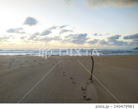Serene coastal landscape at sunrise with footprints leading to a single upright stick in the sand. This artistic image symbolizes journeys, direction, and quiet natural reflection. Serene coastal landscape at sunrise with footprints leading to a single upright stick in the sand. This artistic image symbolizes journeys, direction, and quiet natural reflection. 140479961