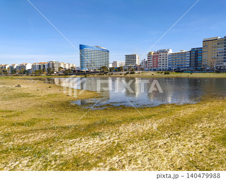 A modern city skyline featuring a curved glass hotel reflecting in a calm coastal lagoon. This vibrant urban landscape is ideal for travel, real estate, and architectural lifestyle projects. A modern city skyline featuring a curved glass hotel reflecting in a calm coastal lagoon. This vibrant urban landscape is ideal for travel, real estate, and architectural lifestyle projects. 140479988