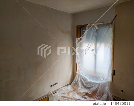 Interior room undergoing renovation with stained plaster walls and a window covered in protective plastic sheeting, capturing the messy preparation phase of home improvement projects. Interior room undergoing renovation with stained plaster walls and a window covered in protective plastic sheeting, capturing the messy preparation phase of home improvement projects. 140480011