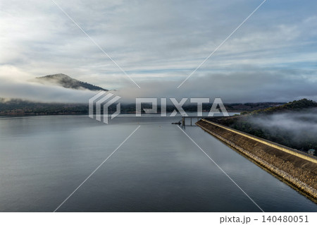 Aerial view of the Glenbawn Dam wall featuring the water intake system and servicing pontoon with fog floating across the water in NSW Australia. Drone photograph captures atmospheric reservoir 140480051