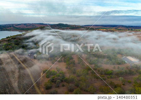 Vertical Aerial image of the Glenbawn Dam wall with morning fog drifting through the surrounding valley in NSW Australia. Drone photograph captures the reservoir, rugged mountain landscape  140480351