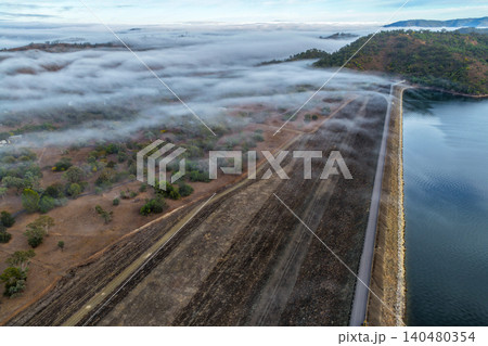 Vertical Aerial image of the Glenbawn Dam wall with morning fog drifting through the surrounding valley in NSW Australia. Drone photograph captures the reservoir, rugged mountain landscape 140480354