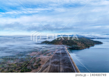 Vertical Aerial view of the Glenbawn Dam wall leading towards the surrounding mountain landscape with fog rolling through the valley in NSW Australia. Drone photograph captures atmospheric low cloud 140480749