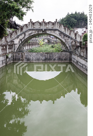 an ornate, weathered stone arch bridge adorned with intricate bas-relief carvings spans gracefully over a canal, casting a flawless, semi-circular reflection onto the calm, greenish waters below. an ornate, weathered stone arch bridge adorned with intricate bas-relief carvings spans gracefully over a canal, casting a flawless, semi-circular reflection onto the calm, greenish waters below. 140481029