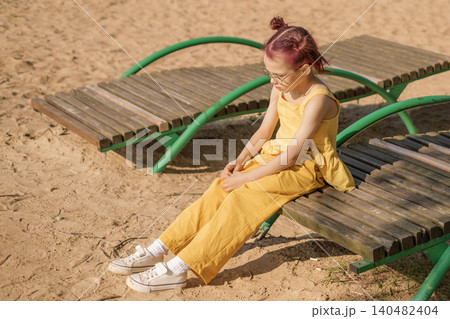 A little lonely sad girl is sitting on a bench on the beach. Child bullying in society A little lonely sad girl is sitting on a bench on the beach. Child bullying in society 140482404