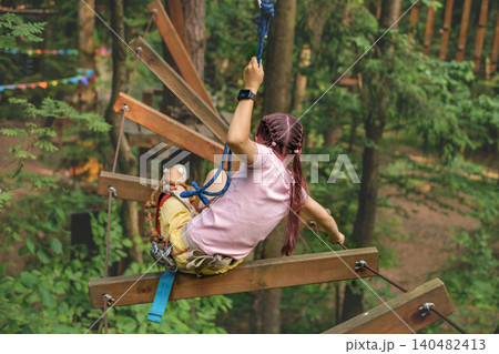 The girl barely passes an obstacle on the suspended bridge of a rope route in an active recreation park The girl barely passes an obstacle on the suspended bridge of a rope route in an active recreation park 140482413