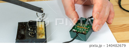 A man works on fixing a broken Christmas tree garland by soldering wires together. He uses tools and parts on a wooden table, focusing on the repair process during the holiday season, banner A man works on fixing a broken Christmas tree garland by soldering wires together. He uses tools and parts on a wooden table, focusing on the repair process during the holiday season, banner 140482679