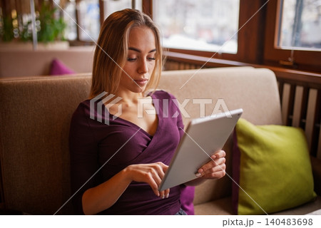A focused blonde woman using a digital tablet while sitting on a cozy sofa inside a cafe 140483698