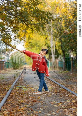 A happy young girl plays with a long wooden stick while standing under autumn trees near a railway track. A happy young girl plays with a long wooden stick while standing under autumn trees near a railway track. 140483738