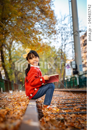 On an autumn day, a little Asian girl holding a book sits on the quaint railway tracks, smiling at the world around her. 140483742