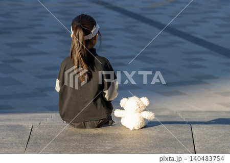 Sad and lonely little girl crying and hugging teddy bear standing by the wall outdoors Sad and lonely little girl crying and hugging teddy bear standing by the wall outdoors 140483754