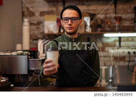A male barista standing behind a bar counter holding a paper coffee cup A male barista standing behind a bar counter holding a paper coffee cup 140483943
