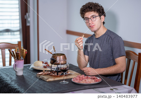 Young man sitting at a table enjoying burgundian fondue with boiling oil, skewering and eating meat, glass of red wine nearby in a cozy apartment dining scene Young man sitting at a table enjoying burgundian fondue with boiling oil, skewering and eating meat, glass of red wine nearby in a cozy apartment dining scene 140483977