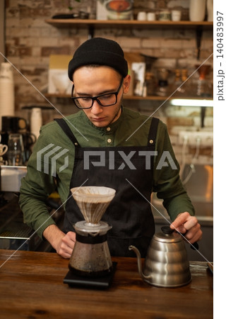 A focused young barista preparing alternative filter coffee with a kettle and a dripper A focused young barista preparing alternative filter coffee with a kettle and a dripper 140483997