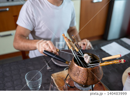 Man cooks beef pieces in a bubbling oil fondue pot at the table, enjoying a social, gourmet dining experience with friends or family in a cozy indoor setting 140483999