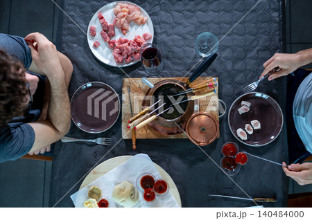 People are sitting at a table sharing a fondue bourguignonne meal, cooking various types of raw meat like beef and chicken in boiling oil, with different sauces and wine for a social dinner 140484000