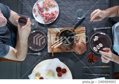 Friends sitting around a table having fondue bourguignonne dinner, holding fondue forks and tasting wine while cooking small pieces of chicken and sausage meat in boiling oil 140484001
