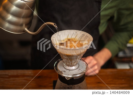 A close up of a barista pouring hot water from a gooseneck kettle into a coffee filter 140484009