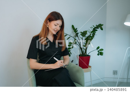 young woman dressed in black medical suit holding a clipboard seated on armchair in modern clinic 140485891