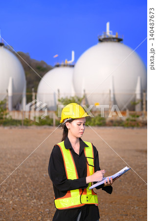 Female industrial engineer checking inspection documents near gas storage tanks at energy facility 140487673
