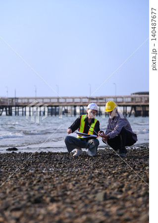 Two engineers stand on a rocky shoreline. They review data on tablets and devices. Pier into calm waters behind them 140487677