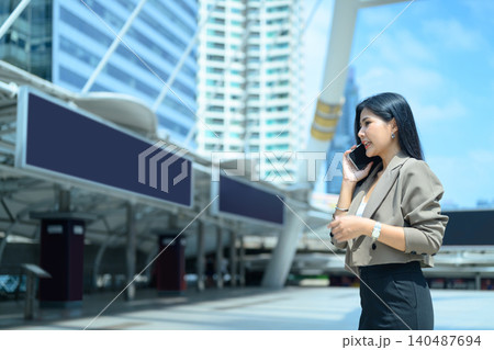 Smiling Asian business woman entrepreneur wearing suit talking with mobile phone standing in city metro 140487694