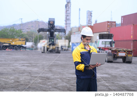 Industrial freight operator managing cargo transport operations with laptop at shipping container yard 140487708