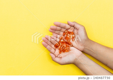 Hands cradle glowing orange capsules against bright yellow background. Soft lighting highlights each pill's glossy texture. Warm tone suggests health, wellness, or self-care 140487730