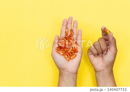 Hand holds orange capsules against a bright yellow background. Fingers gently cradle the pills, suggesting care. Color contrasts emphasize health, wellness, or nutrition. Ideal for supplements 140487732