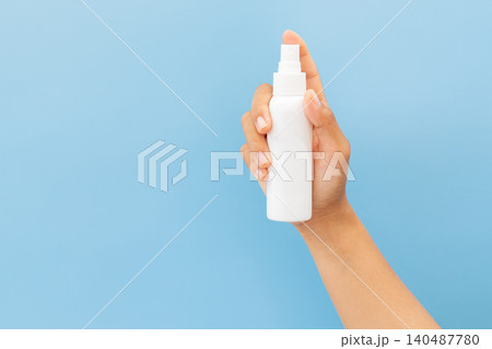 Hand holds white spray bottle against soft blue background. Fingers grip bottle gently, showcasing clean, minimal design. Slight angle highlights spray nozzle and smooth surface 140487780