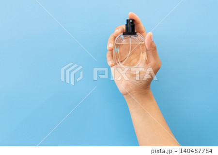 Hand holds clear perfume bottle against blue backdrop. Fingertips cradle glass, black nozzle visible. Simple, clean, minimalist aesthetic dominates frame. Ideal for beauty, lifestyle 140487784
