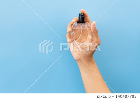 Hand holds clear perfume bottle against blue backdrop. Fingertips cradle glass, black nozzle visible. Simple, clean, minimalist aesthetic dominates frame. Ideal for beauty, lifestyle 140487785