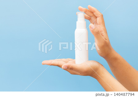Hand holds white spray bottle against soft blue background. Fingers grip bottle gently, showcasing clean, minimal design. Slight angle highlights spray nozzle and smooth surface 140487794