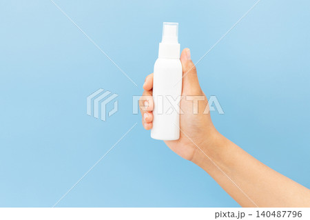 Hand holds white spray bottle against soft blue background. Fingers grip bottle gently, showcasing clean, minimal design. Slight angle highlights spray nozzle and smooth surface 140487796