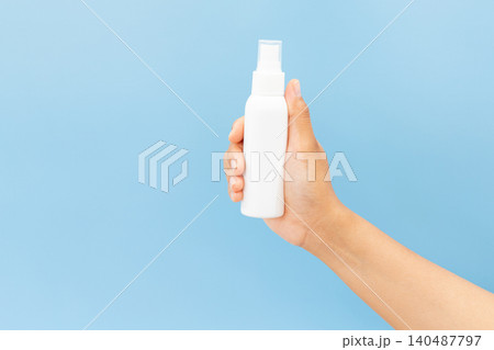 Hand holds white spray bottle against soft blue background. Fingers grip bottle gently, showcasing clean, minimal design. Slight angle highlights spray nozzle and smooth surface 140487797