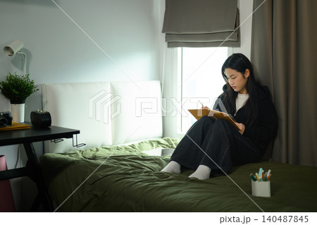 Young Asian Female Student Writing in Notebook on Bed in Cozy University Dorm Room by Window Young Asian Female Student Writing in Notebook on Bed in Cozy University Dorm Room by Window 140487845