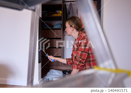 Woman applies dark paint to wall with roller, with ladder blurred in foreground Woman applies dark paint to wall with roller, with ladder blurred in foreground 140491571