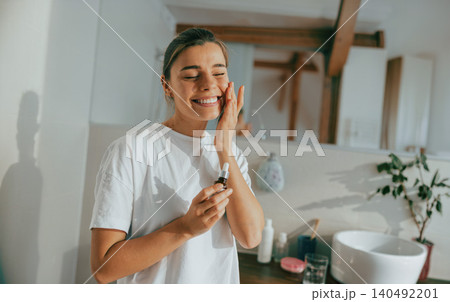 Happy young Caucasian woman applying cosmetic oil serum on face in bright sunny bathroom Happy young Caucasian woman applying cosmetic oil serum on face in bright sunny bathroom 140492201