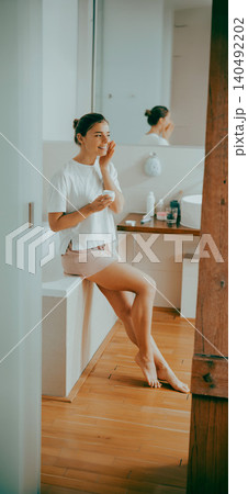 Caucasian young adult woman applying moisturizing cream to her face in a modern bathroom 140492202