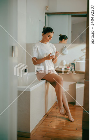 Young Caucasian woman sitting on a bathtub opening cosmetic cream jar in bright bathroom Young Caucasian woman sitting on a bathtub opening cosmetic cream jar in bright bathroom 140492207