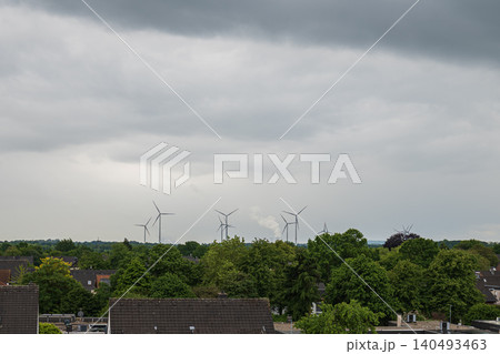 Distant view of wind turbines farm on the horizon behind green trees and house roofs under dark dramatic overcast sky in Germany Distant view of wind turbines farm on the horizon behind green trees and house roofs under dark dramatic overcast sky in Germany 140493463