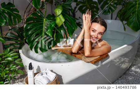 Happy young Caucasian woman relaxing in a bubble bath inside a modern plant filled home bathroom Happy young Caucasian woman relaxing in a bubble bath inside a modern plant filled home bathroom 140493786