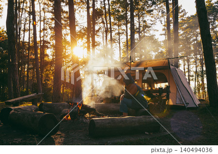 Camper sits by a crackling fire surrounded by logs in a serene forest, with sunlight streaming through the trees and a cozy tent in the background. 140494036