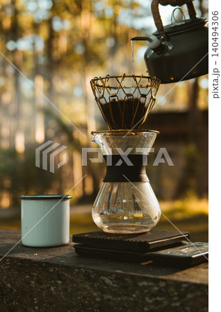 Tranquil outdoor coffee with a kettle pouring water into a pour-over coffee maker on a wooden surface, alongside an enamel mug. Warm sunlight filters through the forest background. Tranquil outdoor coffee with a kettle pouring water into a pour-over coffee maker on a wooden surface, alongside an enamel mug. Warm sunlight filters through the forest background. 140494306